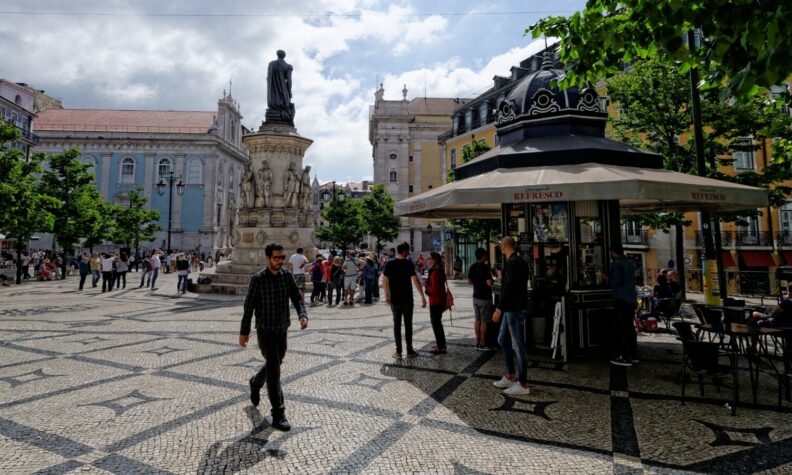 praça da cidade com pessoas a caminhar e um quiosque ao fundo